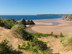 Una playa con dunas de arena, cielo despejado y una costa lejana.