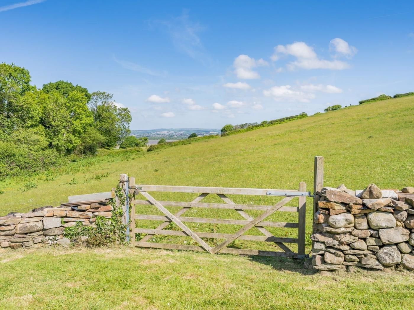 A rural landscape with a wooden gate and stone wall, surrounded by green fields and trees.