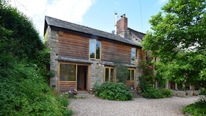 A two-story wooden house with stone accents, a gravel driveway, and lush greenery.