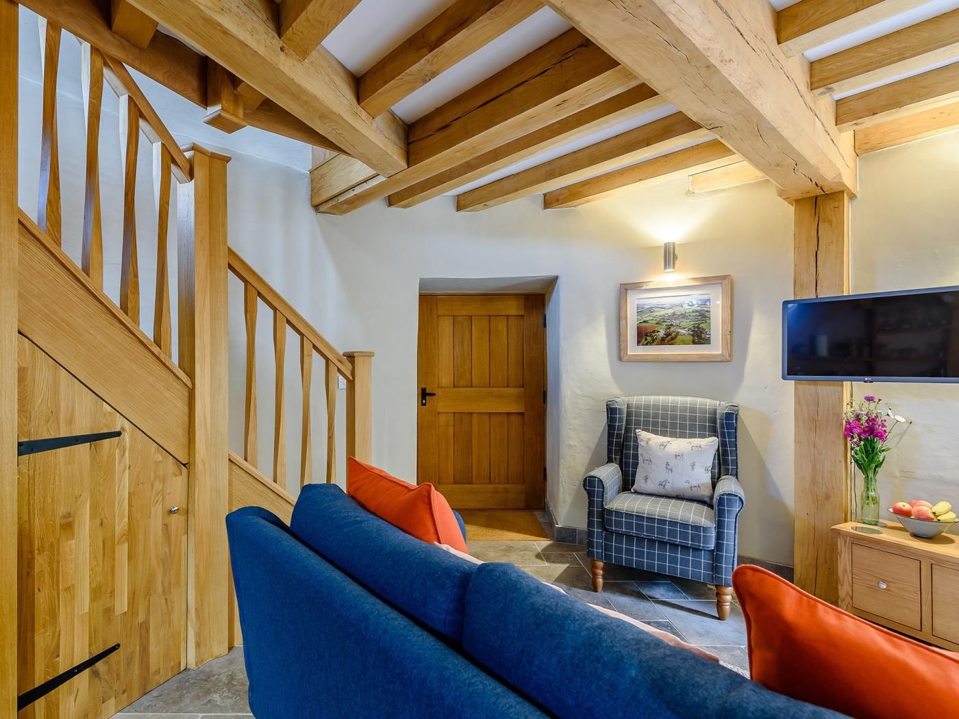 Une pièce avec un escalier en bois, un canapé bleu, un fauteuil à motifs et un téléviseur fixé au mur.