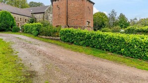 Un bâtiment rural doté d’une tour en briques, entouré de verdure et bordé d’un chemin de gravier.