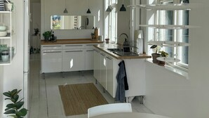 Une cuisine moderne avec des meubles blancs, un plan de travail en bois et un lave-vaisselle encastré. Des plantes en pot sont posées sur le rebord de la fenêtre, et un luminaire suspendu éclaire la pièce.