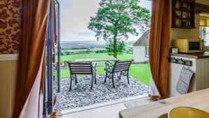 A room with a table and chairs, a view of a tree and fields, and a kitchen area with a microwave. - 1 Bed in Crook (oc-78487) (North Bitchburn)