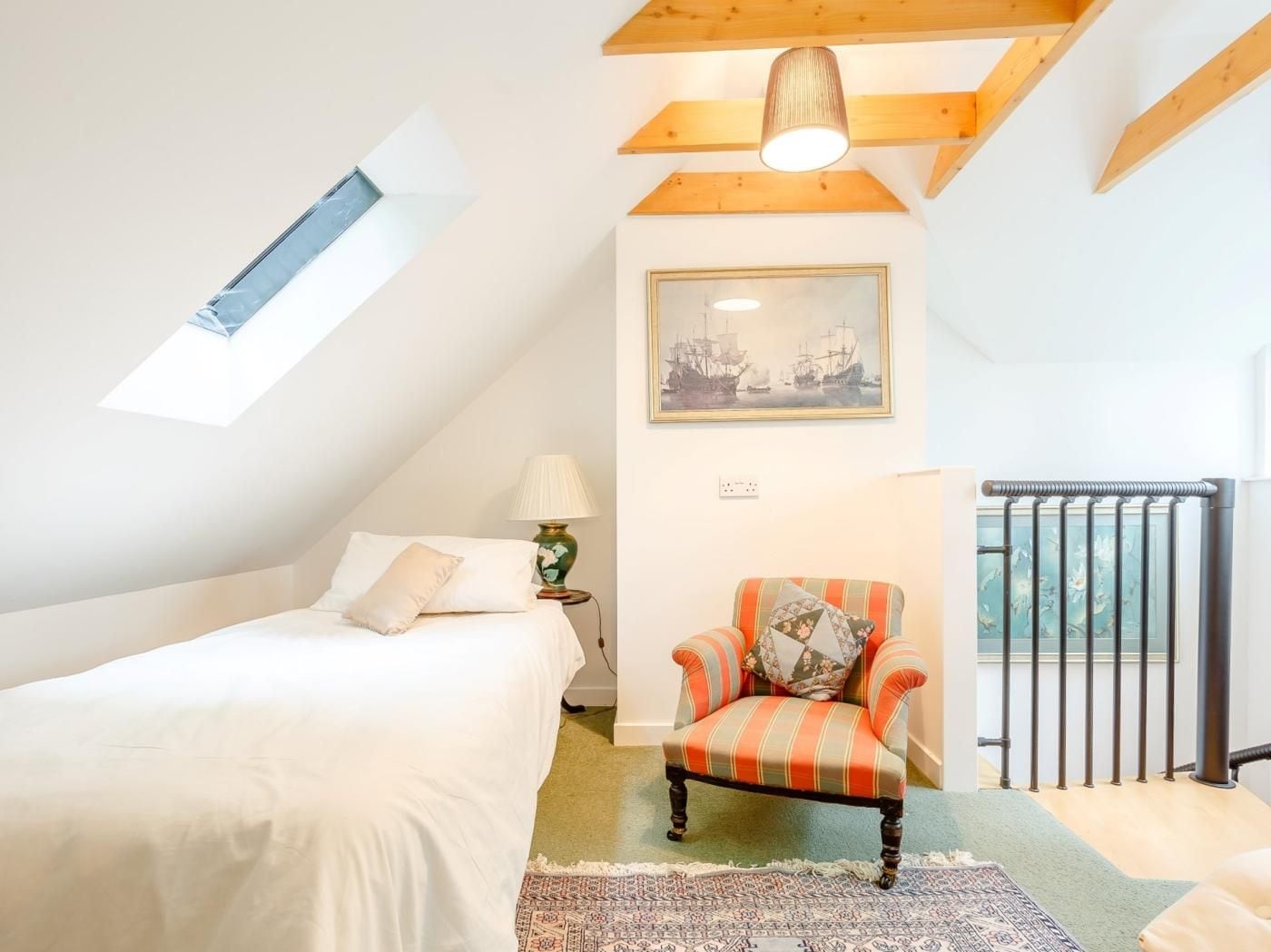 Une chambre avec un plafond en pente, un lit, une chaise, une lampe, un tableau et une vue sur un balcon.