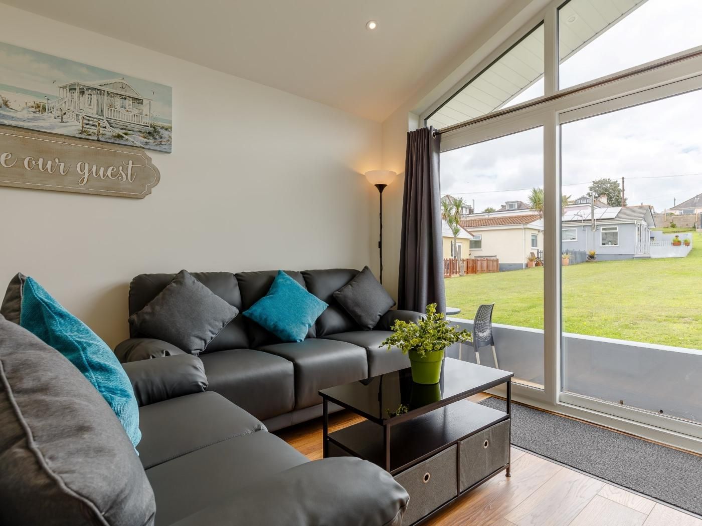 A modern living room with a sofa, a coffee table, and a view of a house through large windows.