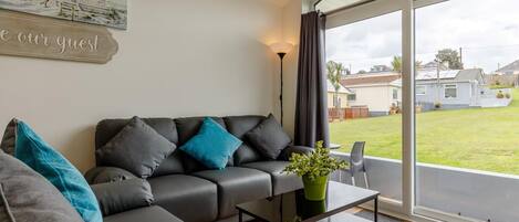 A modern living room with a sofa, a coffee table, and a view of a house through large windows.