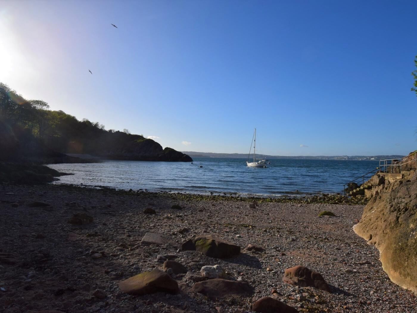 Una platja rocosa amb un veler, cel clar i turons allunyats.