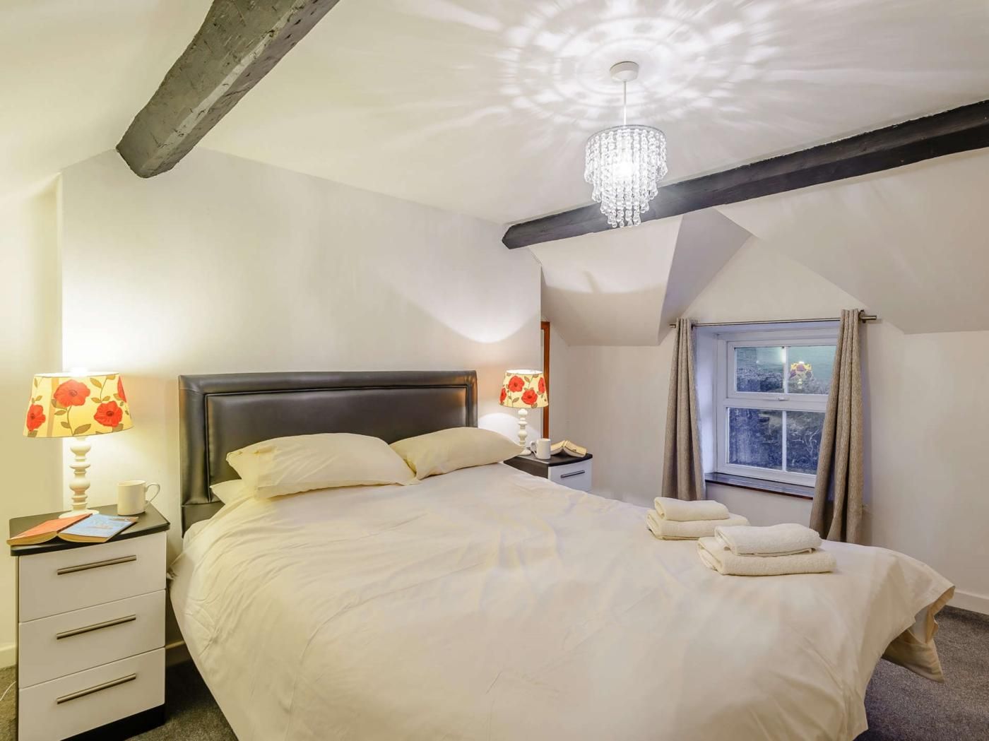 A bedroom with a bed, bedside lamps, a nightstand, a window with curtains, and a chandelier.
