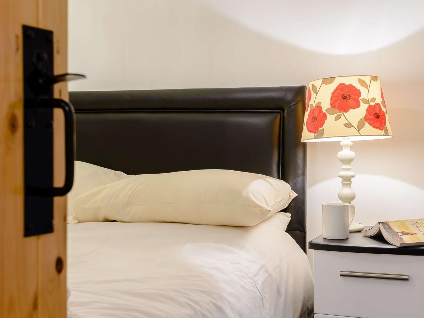 A bed with white bedding, a black headboard, a bedside table with a lamp and a book, and a wooden door.