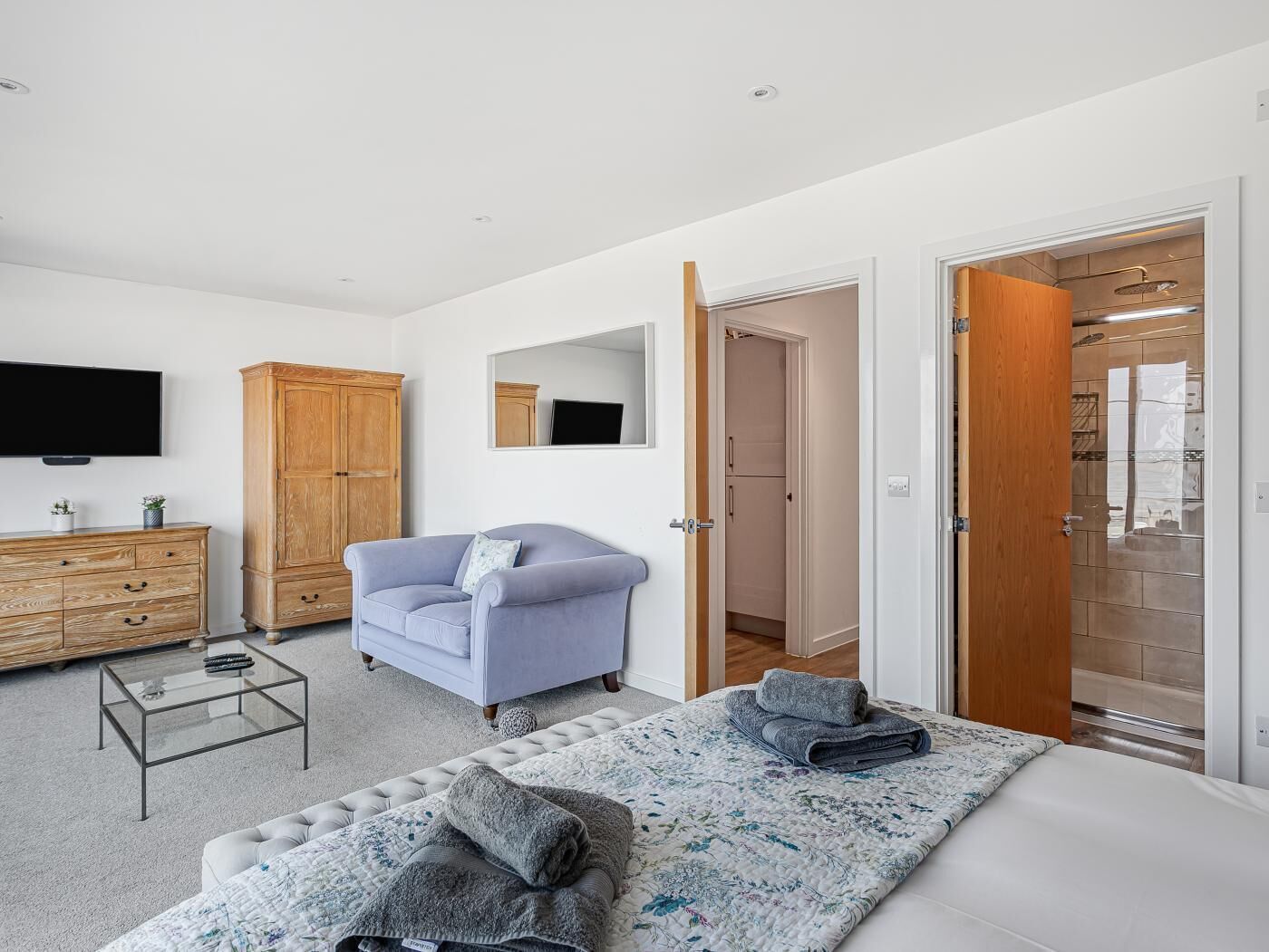 Un dormitorio moderno con cama, sofá, televisor, armario y baño visible a través de una puerta abierta.