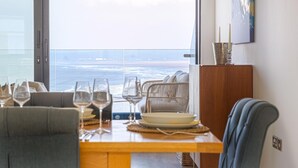 Un coin repas avec vue sur l’océan, une table en bois dressée avec des assiettes et des verres, des chaises en osier et un buffet sur lequel est posé un chandelier.