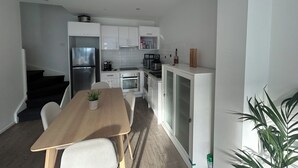 Ett kompakt kök med vita skåp, ett kylskåp, en kaffebryggare och en matplats med ett bord och stolar.