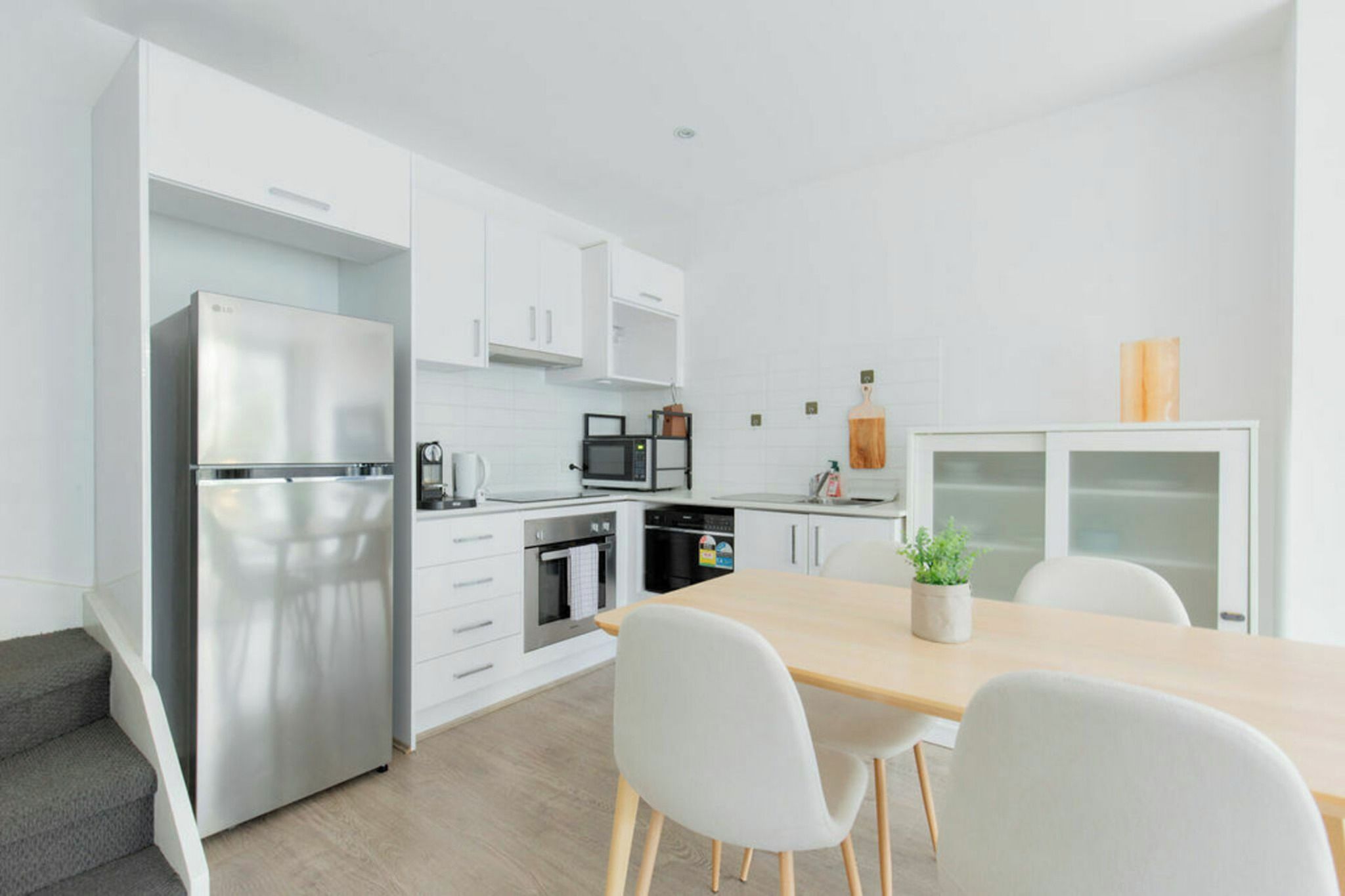 A modern kitchen with white cabinetry, a stainless steel refrigerator, a microwave, and a dining area with a table and chairs.