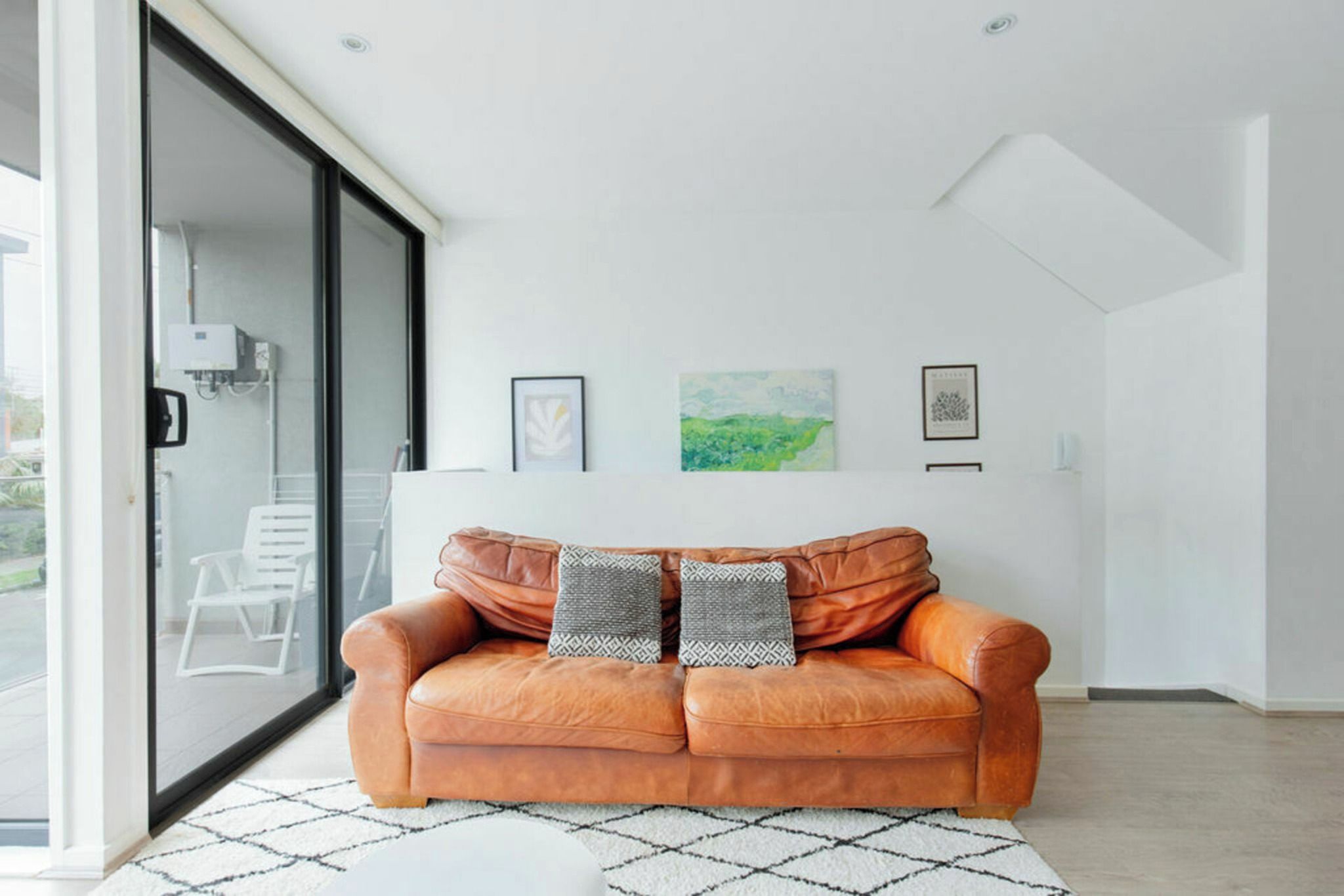 A modern living room with a leather sofa, two patterned cushions, a white rug, and a glass door leading outside.