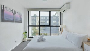 Ett hotellrum med en stor säng, ett fönster med utsikt över byggnader, en krukväxt och en tavla på väggen.