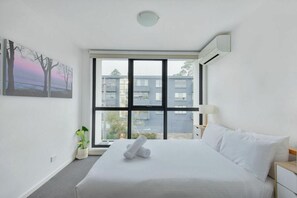 A hotel room with a large bed, a window with a view of buildings, a potted plant, and a painting on the wall. - Beautiful 3BDR 2BA Townhouse in St Kilda East (Saint Kilda East)
