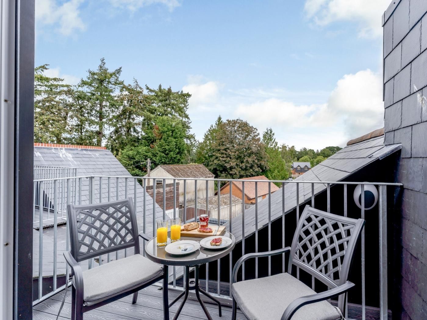 A balcony with two chairs and a table, a breakfast tray with toast and juice, and a view of neighboring houses and trees.