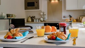 Aamiaistarjoilu, jossa on croissantteja, mansikoita, mustikoita, appelsiinimehua ja purkki hilloa keittiön työtasolla.