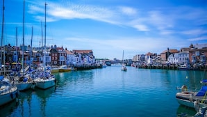 A marina with boats docked, a bridge in the distance, and buildings along the water.