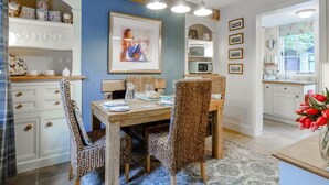 Un comedor con una mesa de madera, sillas de mimbre, un tapete azul y una cocina al fondo.