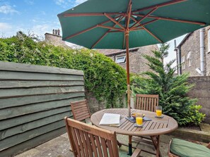 An outdoor patio with a round wooden table, two chairs, a green umbrella, and a glass of orange juice. - 3 Bed in Middleham (oc-78935) (Middleham)