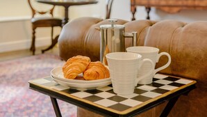 Ein gemütliches Wohnzimmer mit einem braunen Ledersofa, einem kleinen runden Tisch mit karierter Tischdecke, einer Croissantplatte, zwei weißen Tassen und einer silbernen Kaffeekanne.