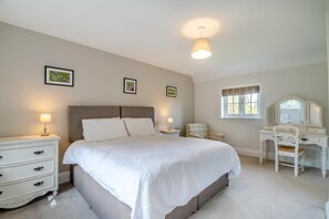 Ein Schlafzimmer mit einem großen Bett, Nachttischen, einem Schminktisch, einem Sessel, einem Schreibtisch mit Spiegel und einem Fenster mit Vorhängen.