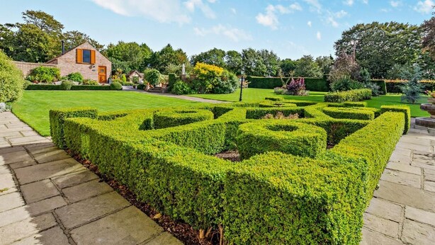 A well-maintained garden with geometric hedge designs, a brick building with a door, and a clear sky. - 2 Bed in Driffield (oc-79280) (Wold Newton)
