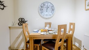 A dining area with a round wall clock, wooden chairs, a table set for breakfast, and a cabinet with a decorative item. - 1 Bed in Lymington (oc-79332) (Milford on Sea)
