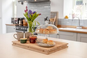 A kitchen counter with a glass display case of scones, a mug, a jar of jam, and a small green bowl. - 6 Bed in Petrockstow (oc-79407) (Petrockstow)