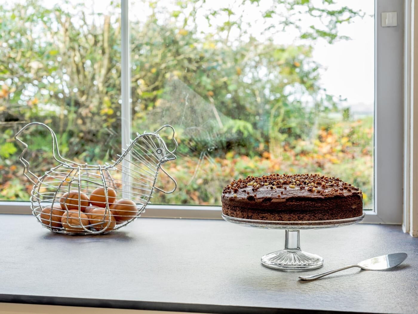 A window with a metal egg holder and a chocolate cake on a glass stand.