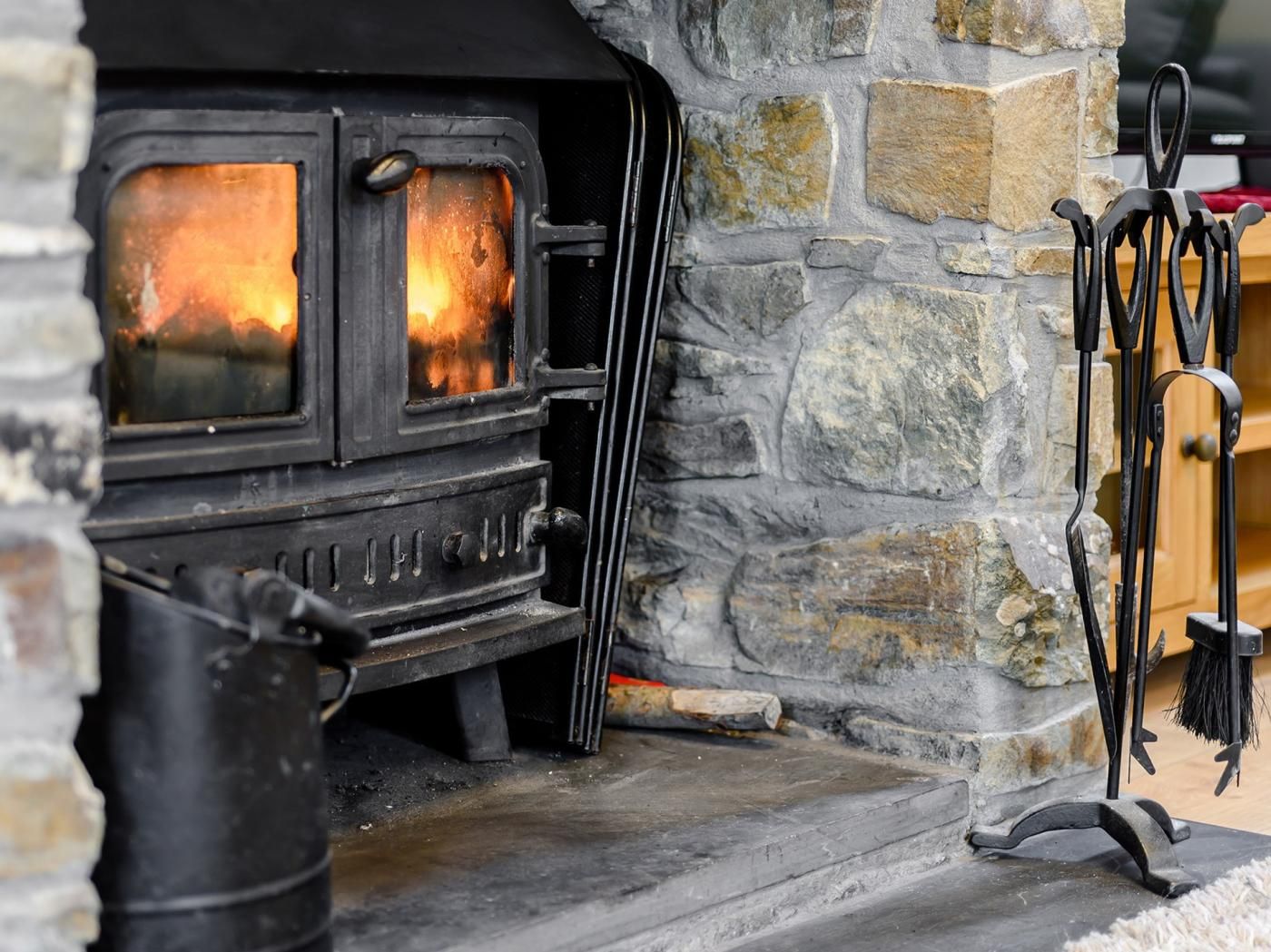 A stone fireplace with a black stove, a chimney, and a set of black tools hanging on the wall.
