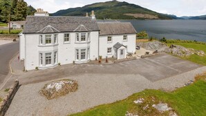 A white two-story house with a driveway, a small garden, and a view of a lake and hills. - 4 Bed in Dunoon (oc-79529) (Ardentinny)