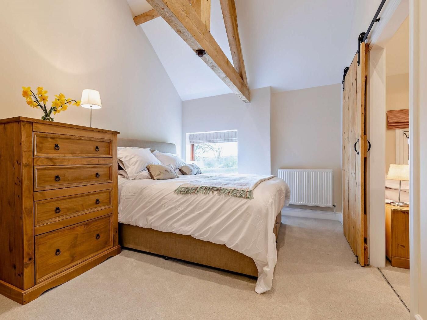 A bedroom with a wooden dresser, a bed with white bedding, a radiator, and a window with a view.