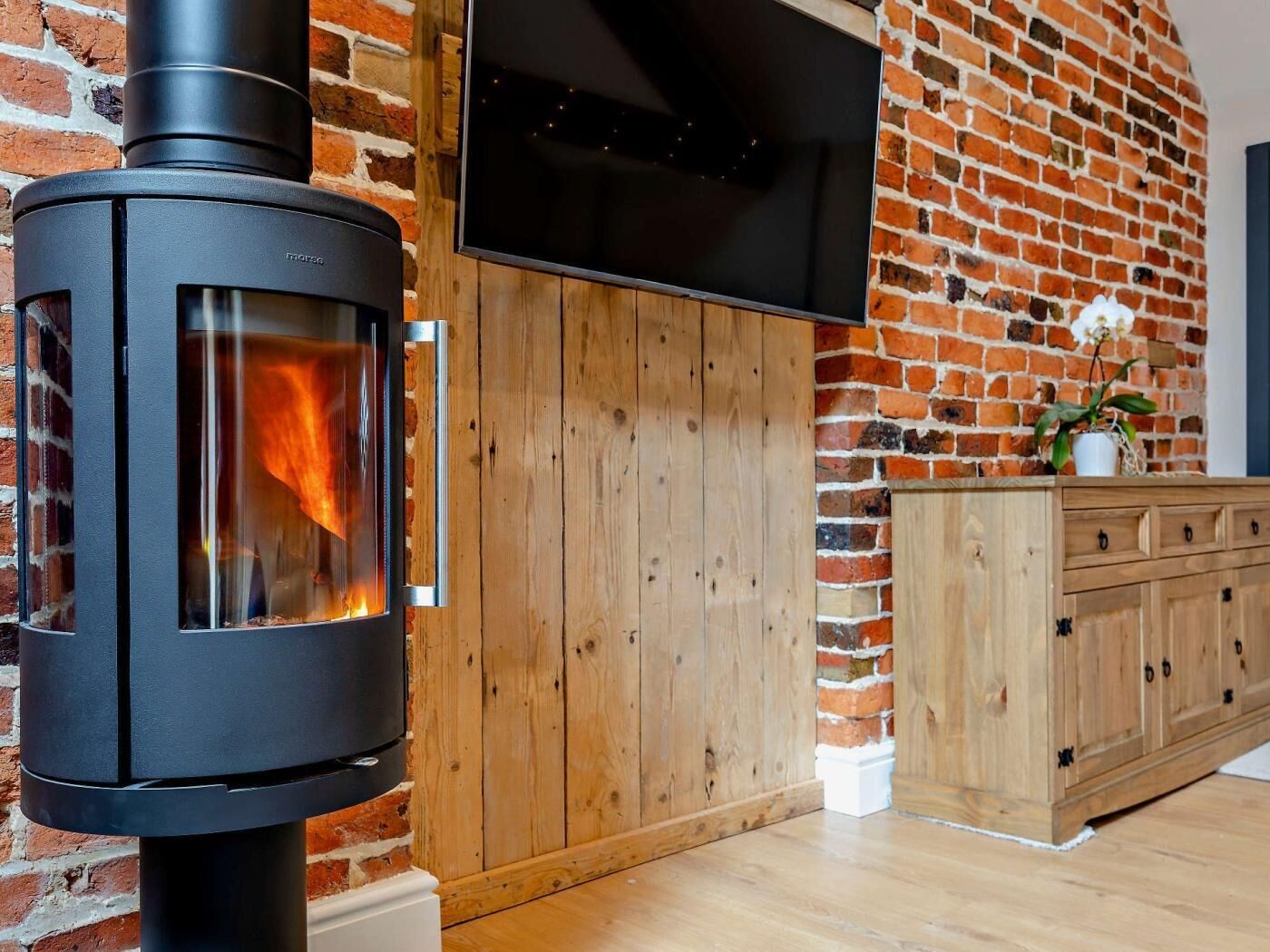 A wood-burning stove with a glass front, a wooden cabinet, and a brick wall with a mounted TV.