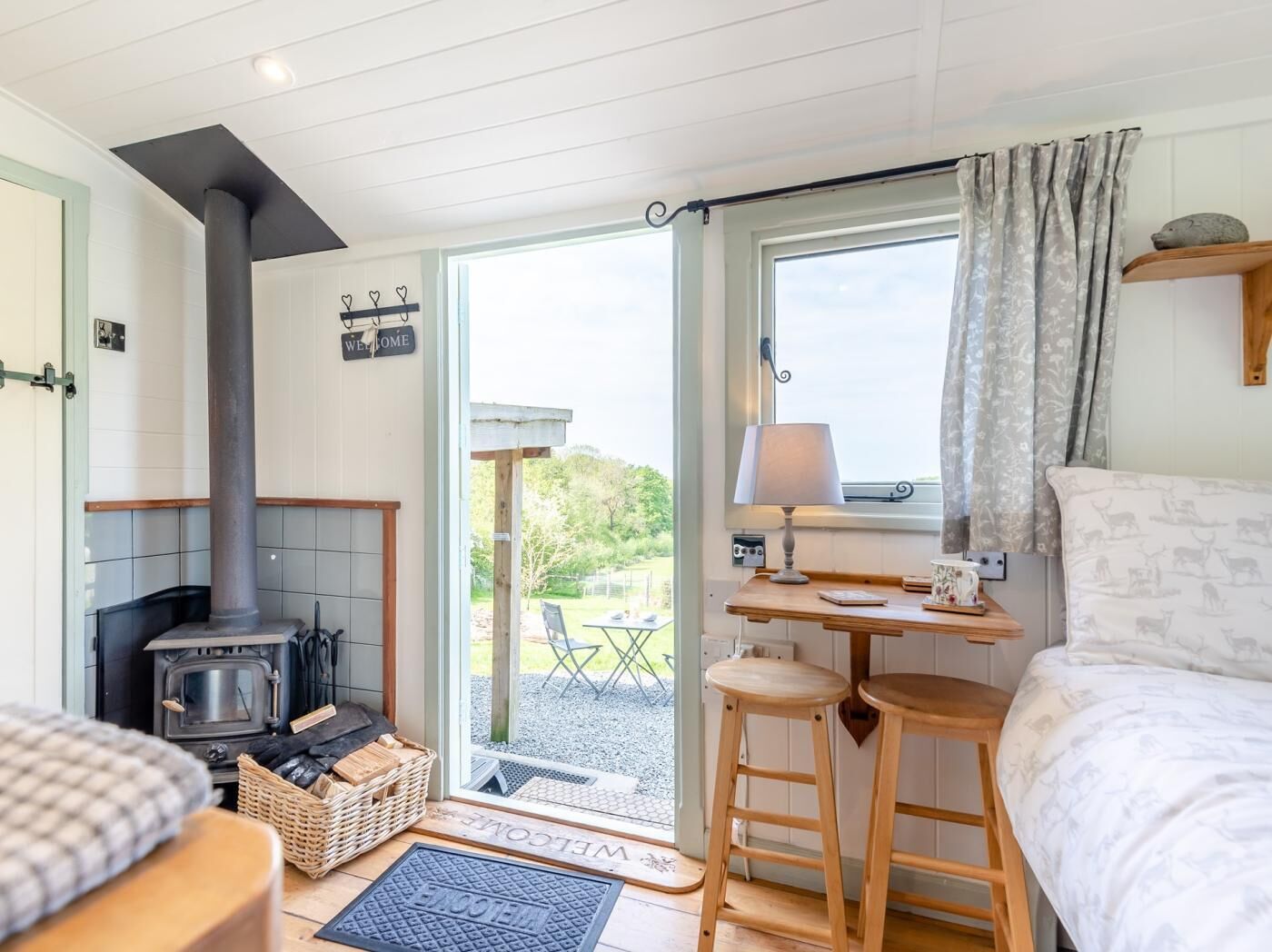 A cozy room with a wood-burning stove, a bed, a table with a lamp, and a view of the outdoors.