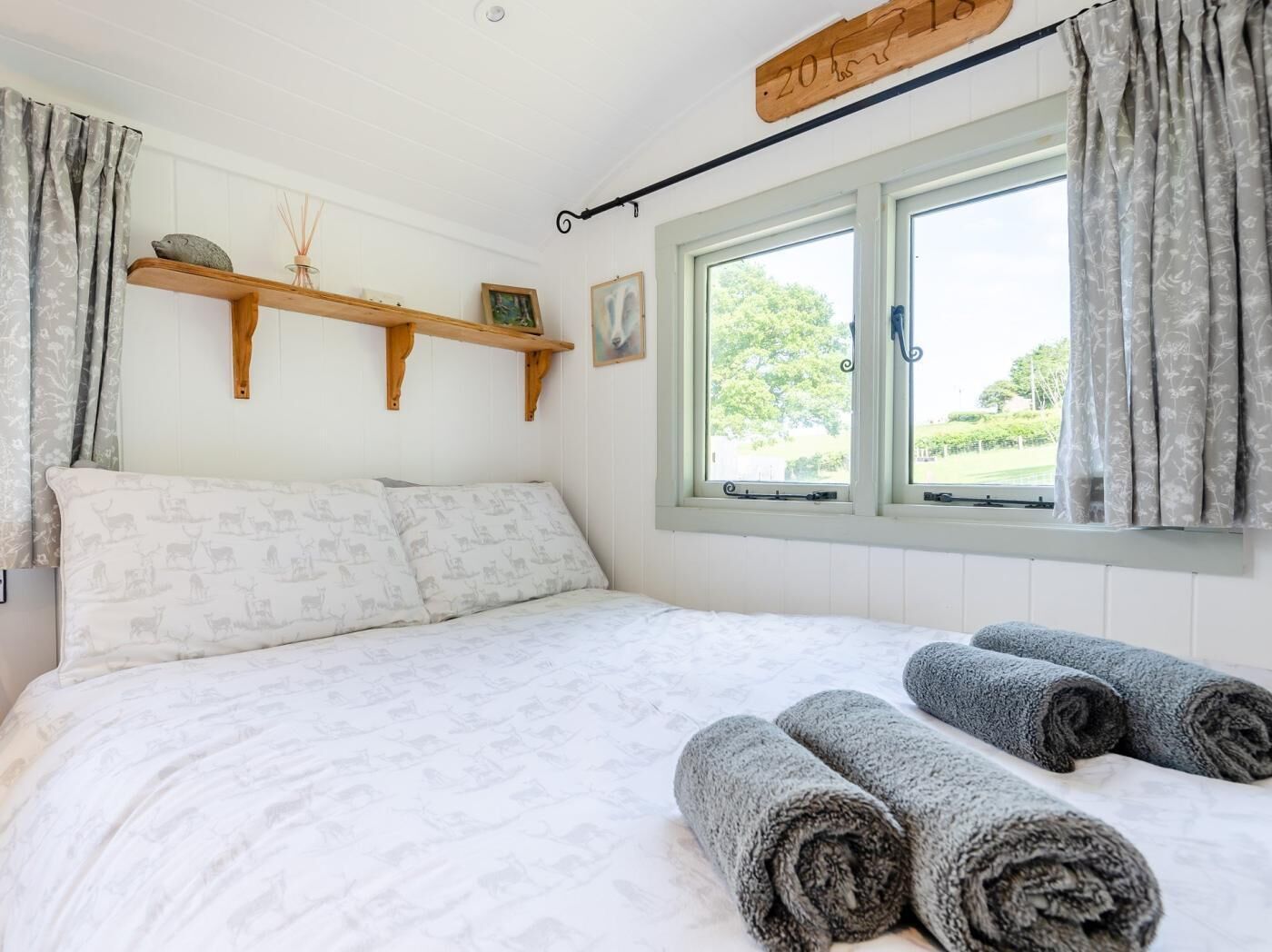 A bedroom with a bed, two windows, a wooden shelf with decorative items, and rolled towels on the bed.