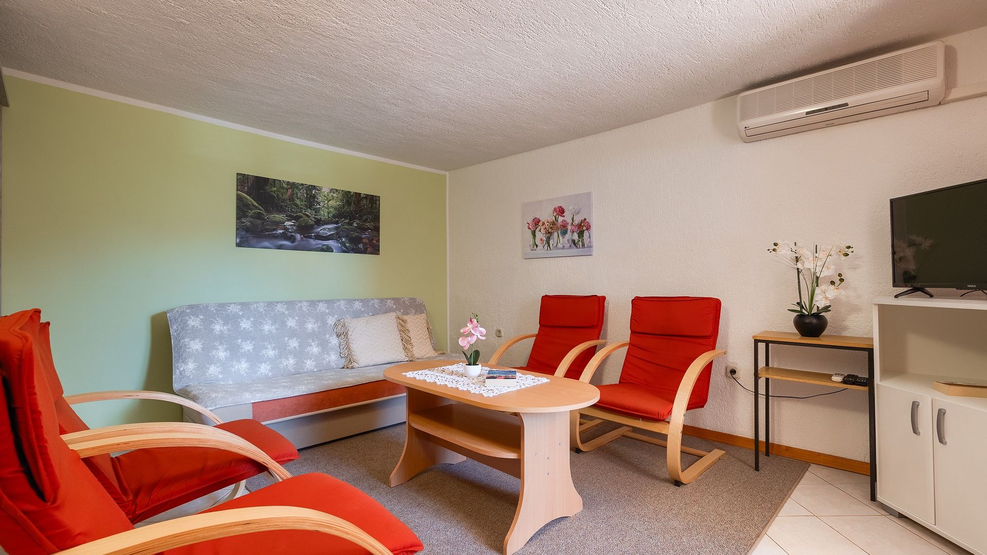 A living room with a sofa, two red chairs, a table with a book and flowers, a TV, and an air conditioner.