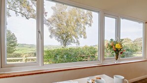 A room with a large window overlooking a rural landscape, a wooden table with a magazine, two white mugs, and a vase with flowers. - 2 Bed in Abergele (oc-80629) (Gwytherin)