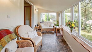 A sunlit room with wicker furniture, a view of the outdoors, and a window with a vase of flowers. - 2 Bed in Abergele (oc-80629) (Gwytherin)
