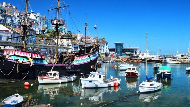 Ein Hafen mit verschiedenen Booten, darunter ein großes Schiff mit bunten Segeln und kleinere, anlegende Boote.