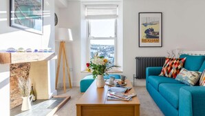 Ein modernes Wohnzimmer mit einem Holztisch, einem blaugrünen Sofa, einem gerahmten Bild und Blick auf die Stadt durch ein Fenster.
