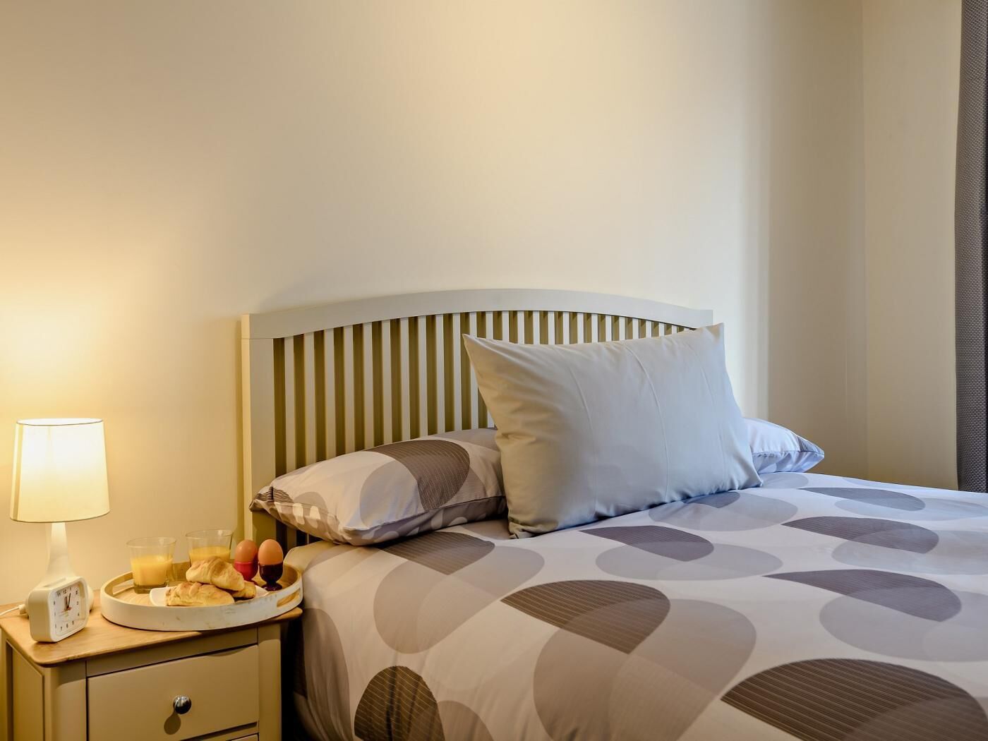 A hotel room with a bed, a bedside table with a lamp, a clock, and a tray with breakfast items.