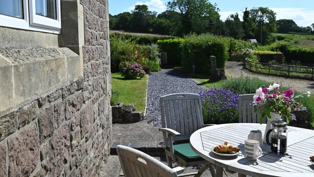A stone building with a window, a paved pathway, and a round wooden table with chairs, a plate of food, and a coffee pot. - 3 Bed in Chepstow (oc-81047) (Shirenewton)