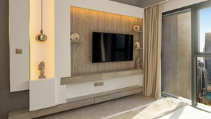 A modern living room with a flat-screen TV mounted on a wooden panel wall, a sculpture on the left, and a view of the city through a large window. - Spacious 3.5BR Burj LED View High Floor  (Dubai)