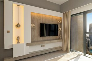 A modern living room with a flat-screen TV mounted on a wooden panel wall, a sculpture on the left, and a view of the city through a large window. - Spacious 3.5BR Burj LED View High Floor  (Dubai)