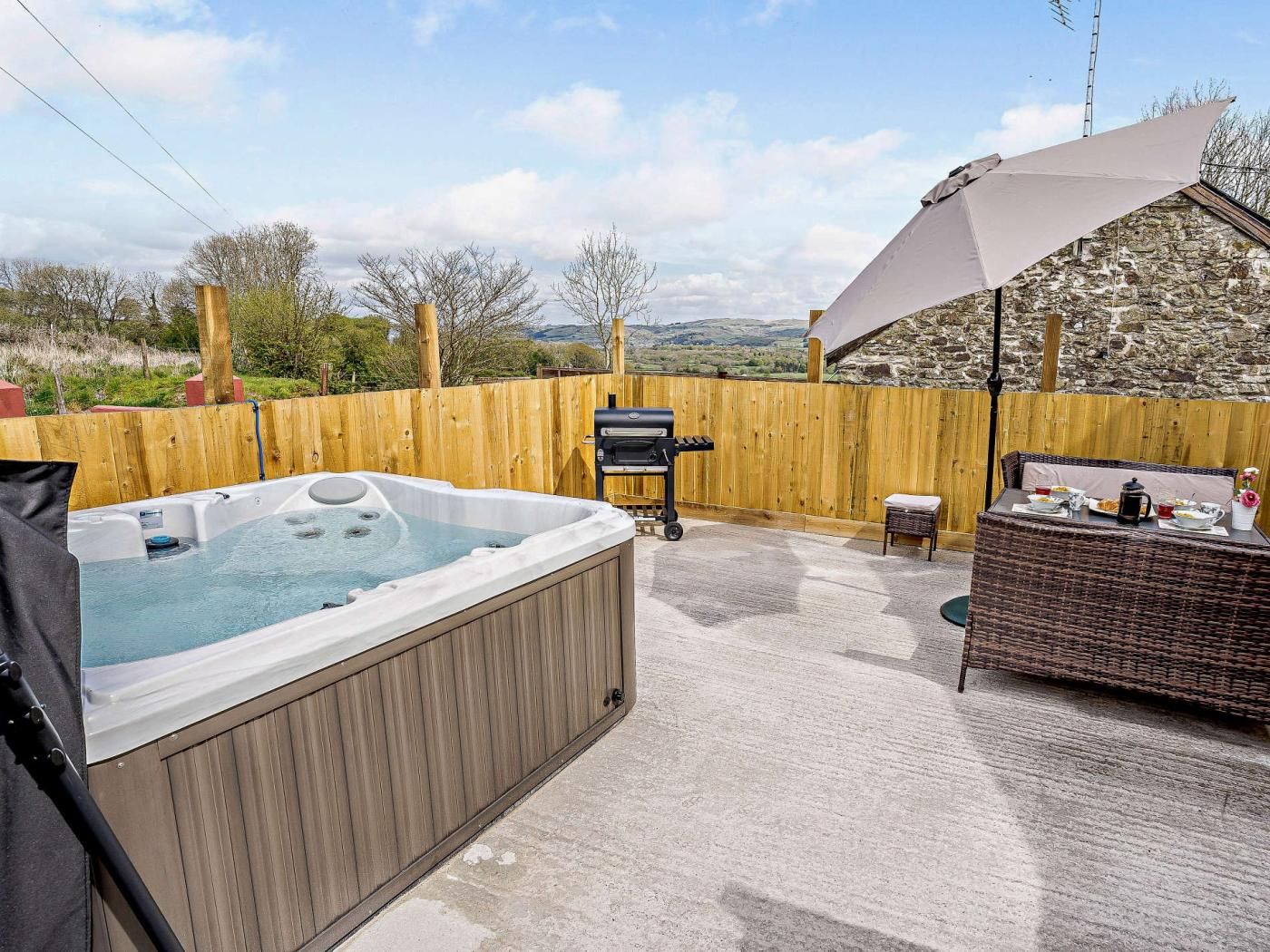 A hot tub, outdoor grill, and wicker furniture on a patio with a view of the countryside.