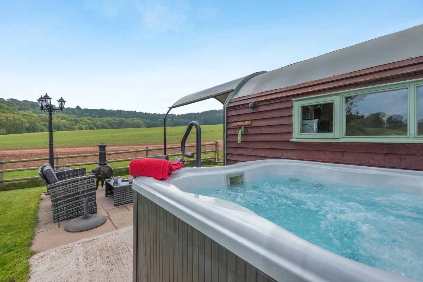 A hot tub with a wooden deck, a window with a view of the countryside, and outdoor furniture.