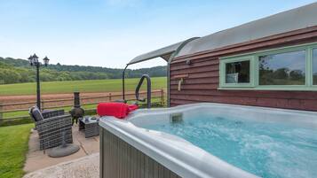 A hot tub with a wooden deck, a window with a view of the countryside, and outdoor furniture.