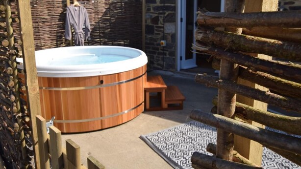 A wooden hot tub in a stone-walled outdoor area with a wooden bench and a patterned rug. - 1 Bed in Launceston (oc-81332) (Saint Giles on the Heath)
